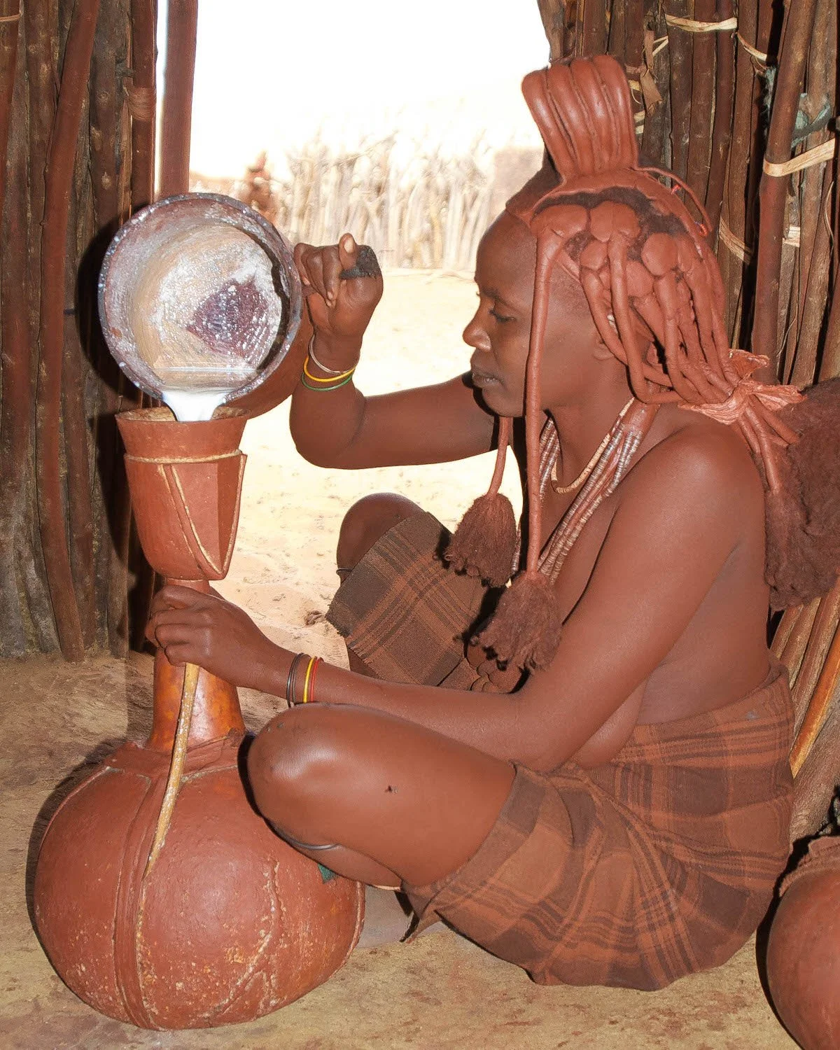 A person in traditional attire, with hairstyle made of braided hair styled to resemble a cooking pot, is sitting inside a wooden hut, pouring water from a clay pot into a wooden container.