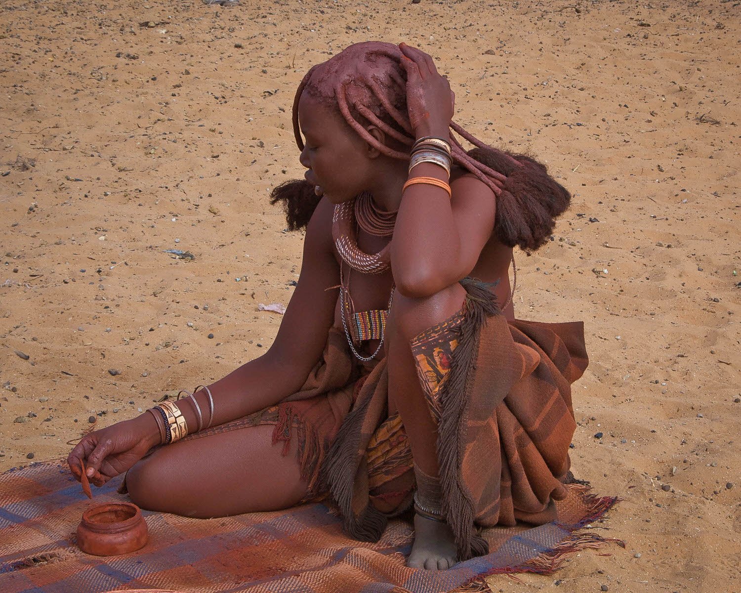 A woman with traditional jewelry and a patterned wrap sitting on a mat on sandy ground, holding her hair with one hand and working with her other hand on a small container.