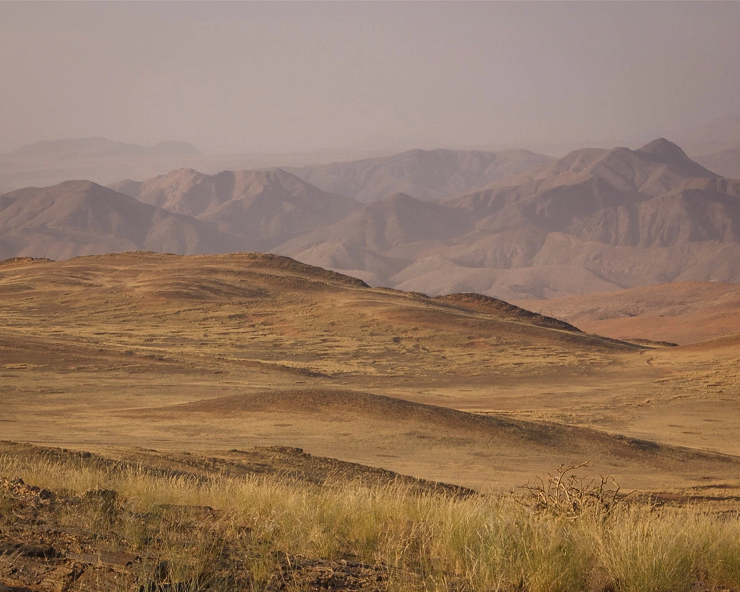A vast desert landscape with rolling hills and distant mountain ranges, featuring sparse vegetation and a hazy sky.