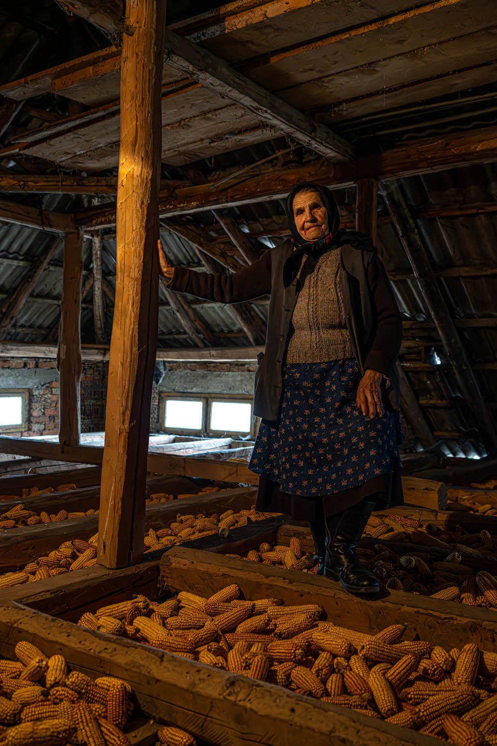 An elderly woman standing inside an attic filled with dried corncobs, holding onto a wooden support beam.