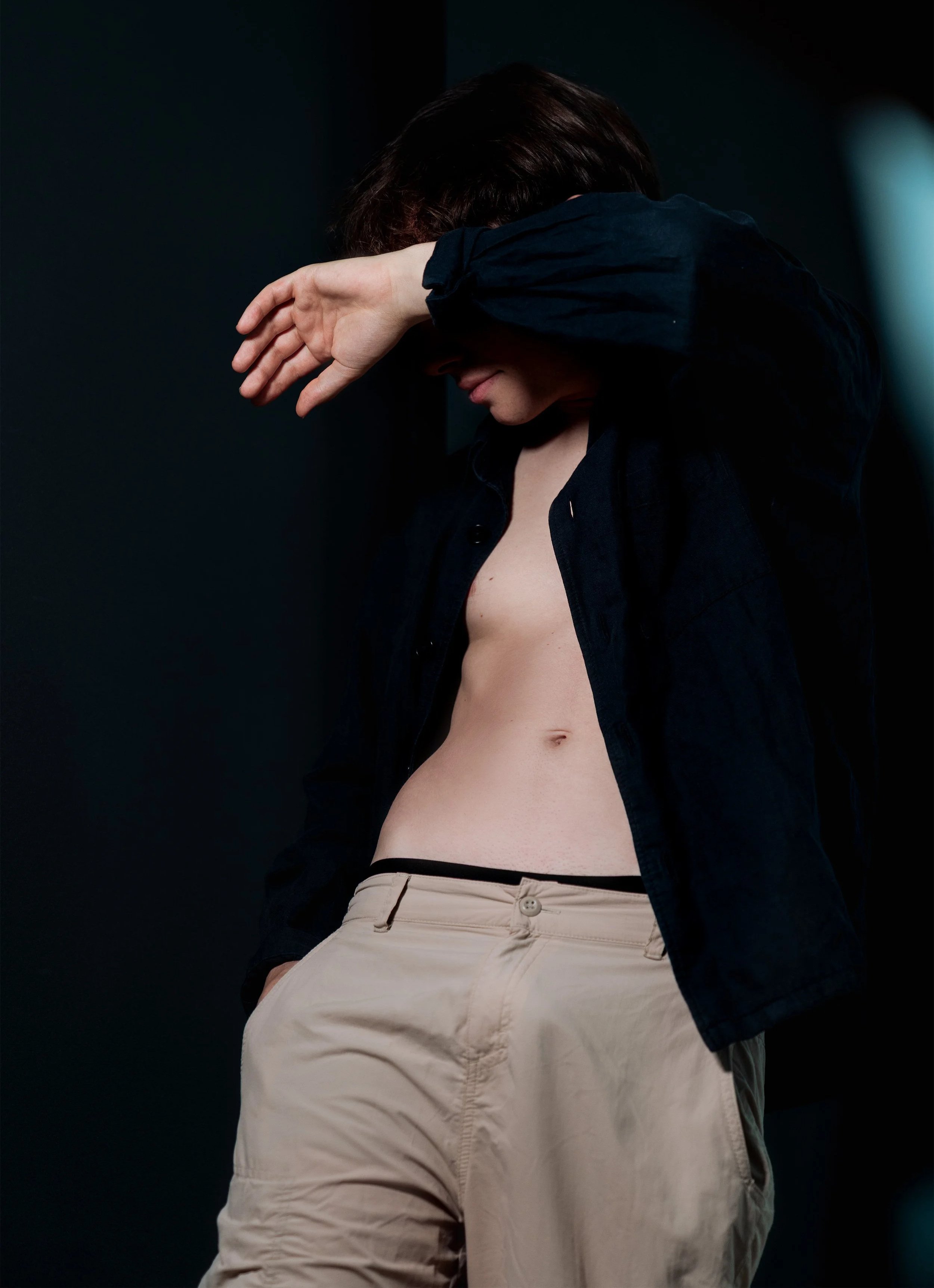 Une personne en tenue décontractée avec une chemise noire et un pantalon beige, qui couvre partiellement son visage avec son bras, dans un environnement sombre.