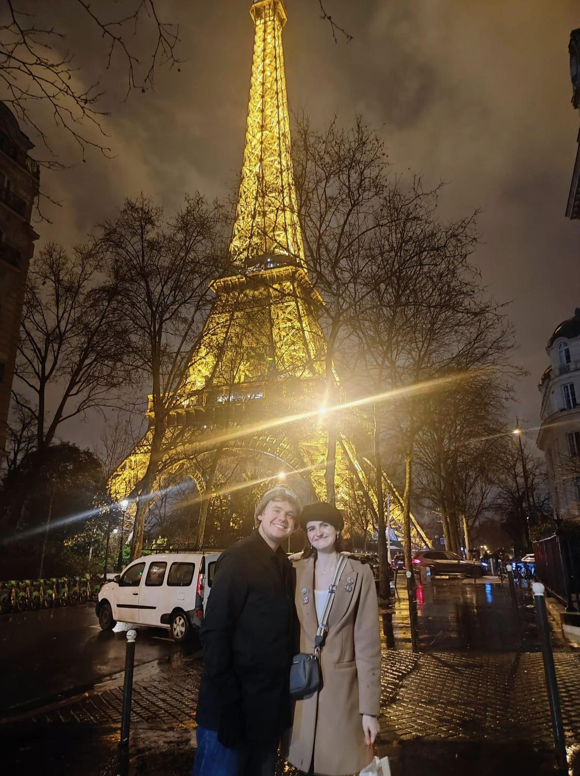 Two people standing together at night with the illuminated Eiffel Tower in the background, surrounded by trees and wet pavement.