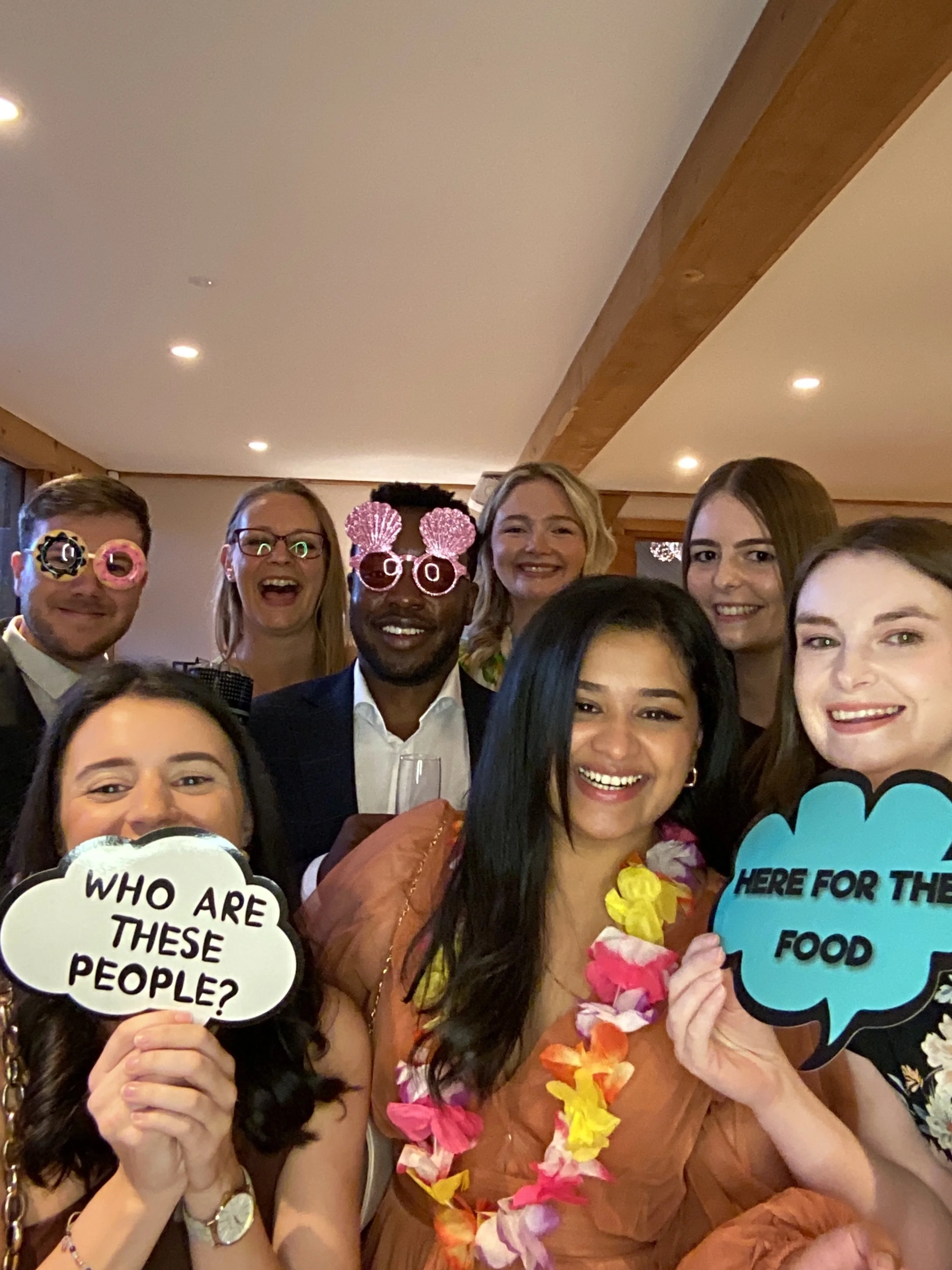Group of smiling people at a party, some wearing novelty glasses, holding signs that say 'Who are these people?' and 'Here for the food,' with a festive, tropical theme.