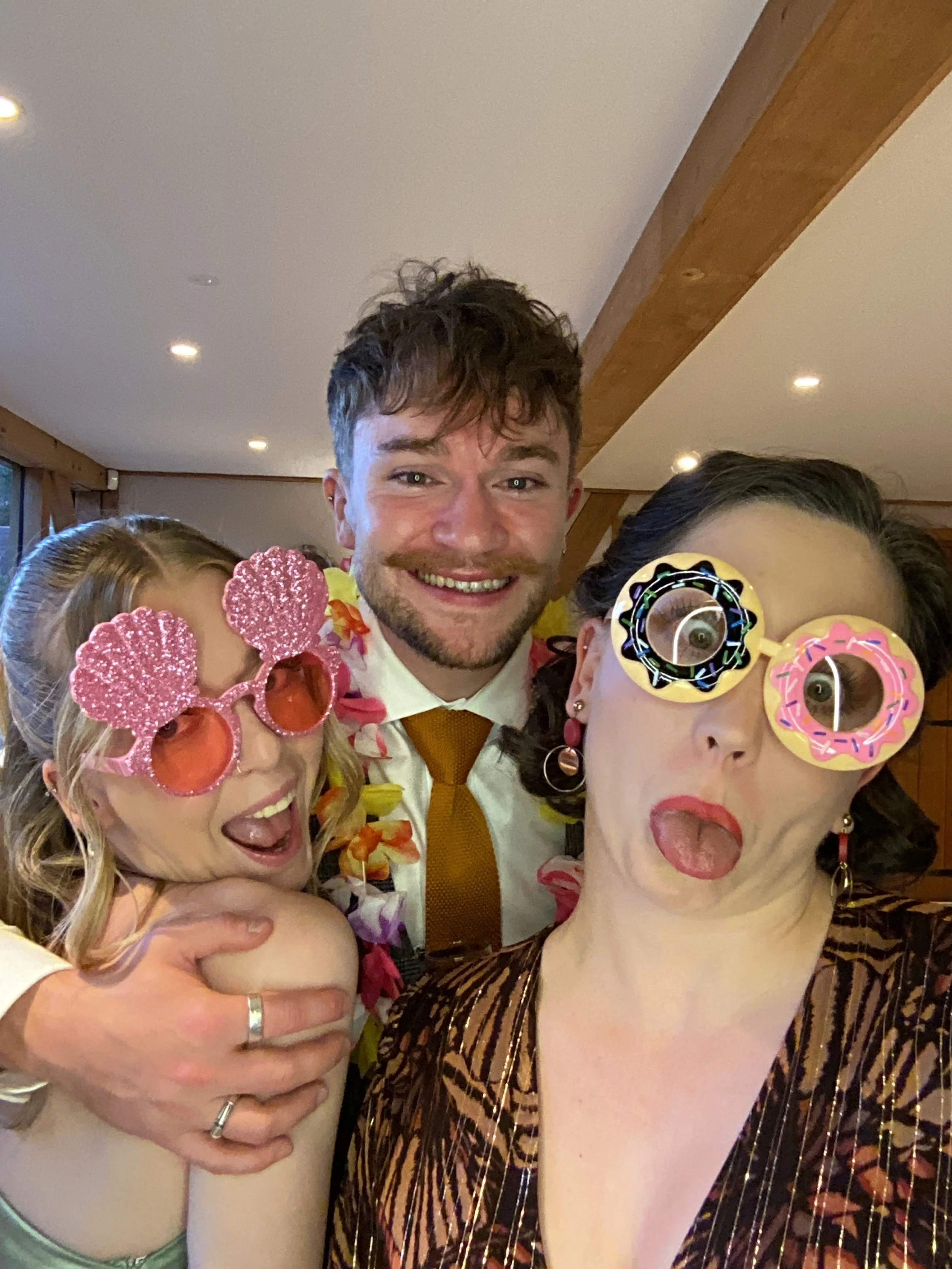 Three people celebrating, wearing colorful glasses and leis, making funny faces indoors.