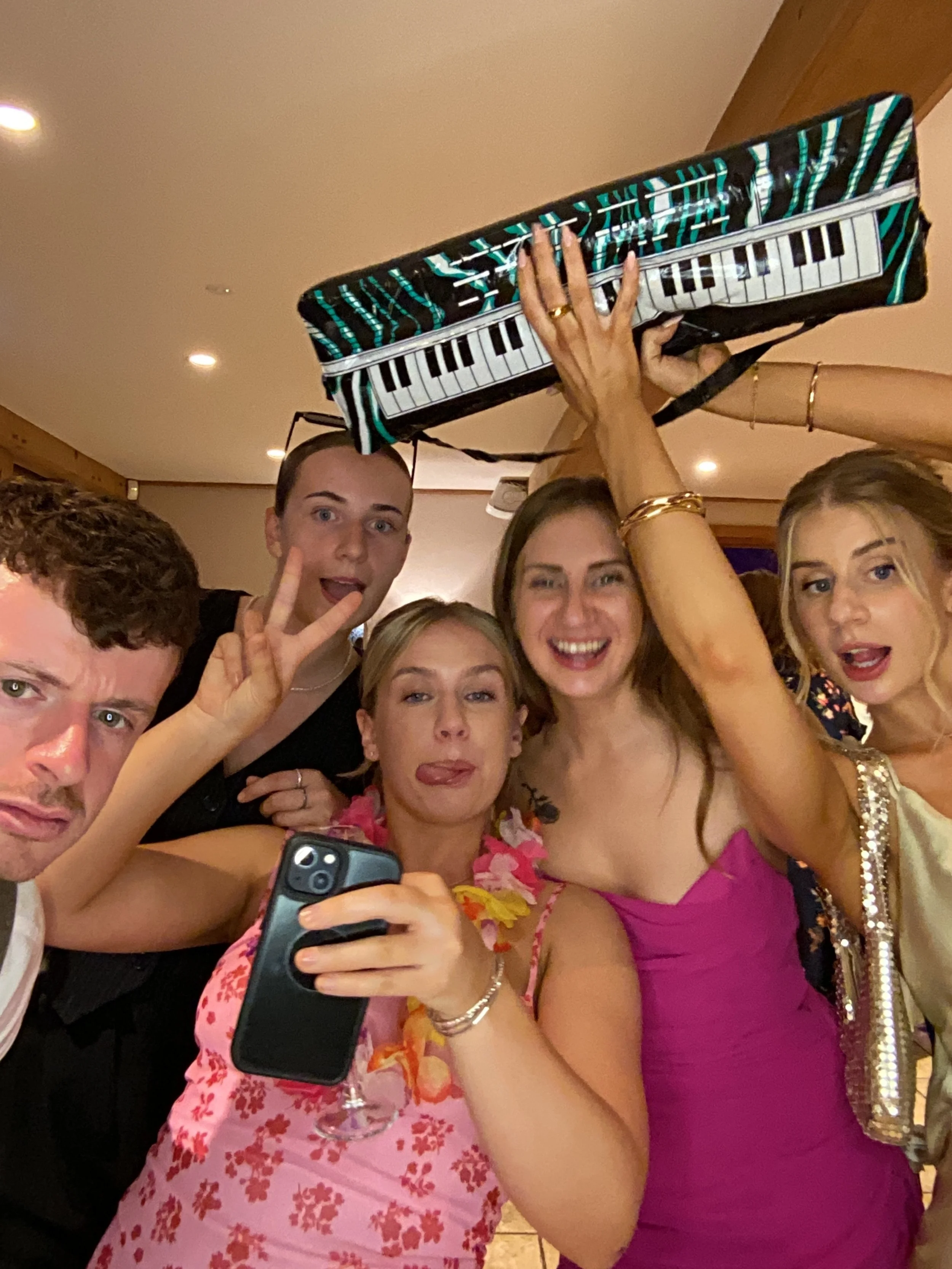 Group of six friends celebrating at a party, one holding a keyboard-shaped purse overhead, making silly faces, with one person taking a selfie, dressed in colorful, festive attire.