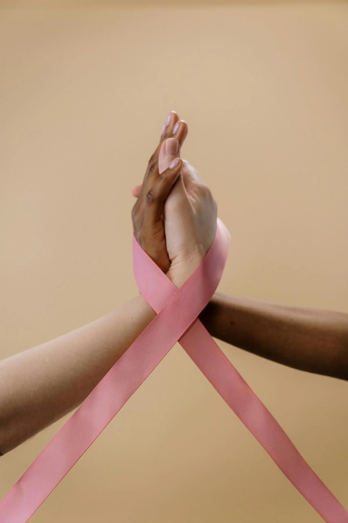 Two hands, one dark-skinned and one light-skinned, clasped together with pink ribbon wrapped around their wrists, against a beige background.