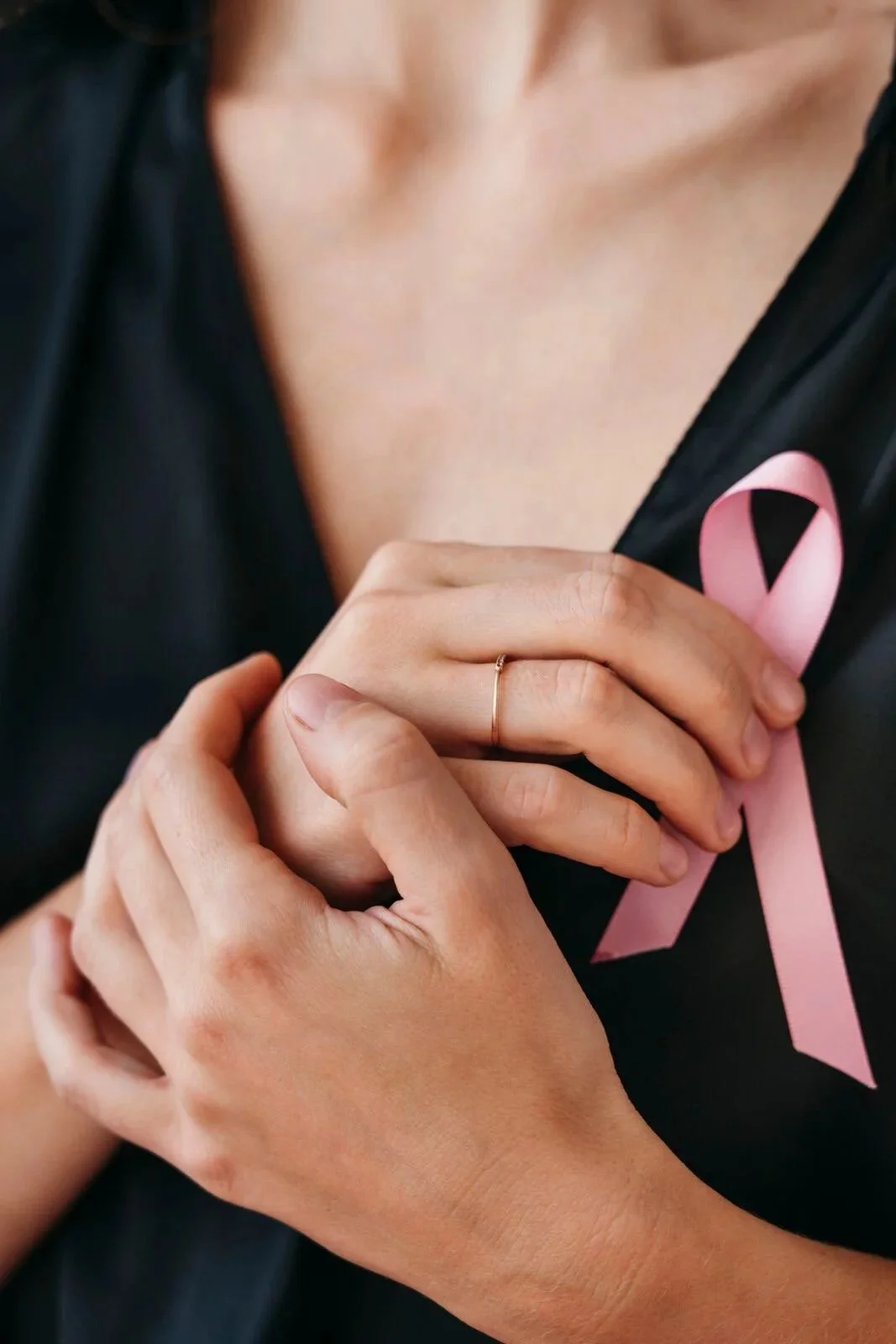 Close-up of a woman's hands holding a pink ribbon pin on her black clothing, symbolizing breast cancer awareness, with a delicate ring on her finger.