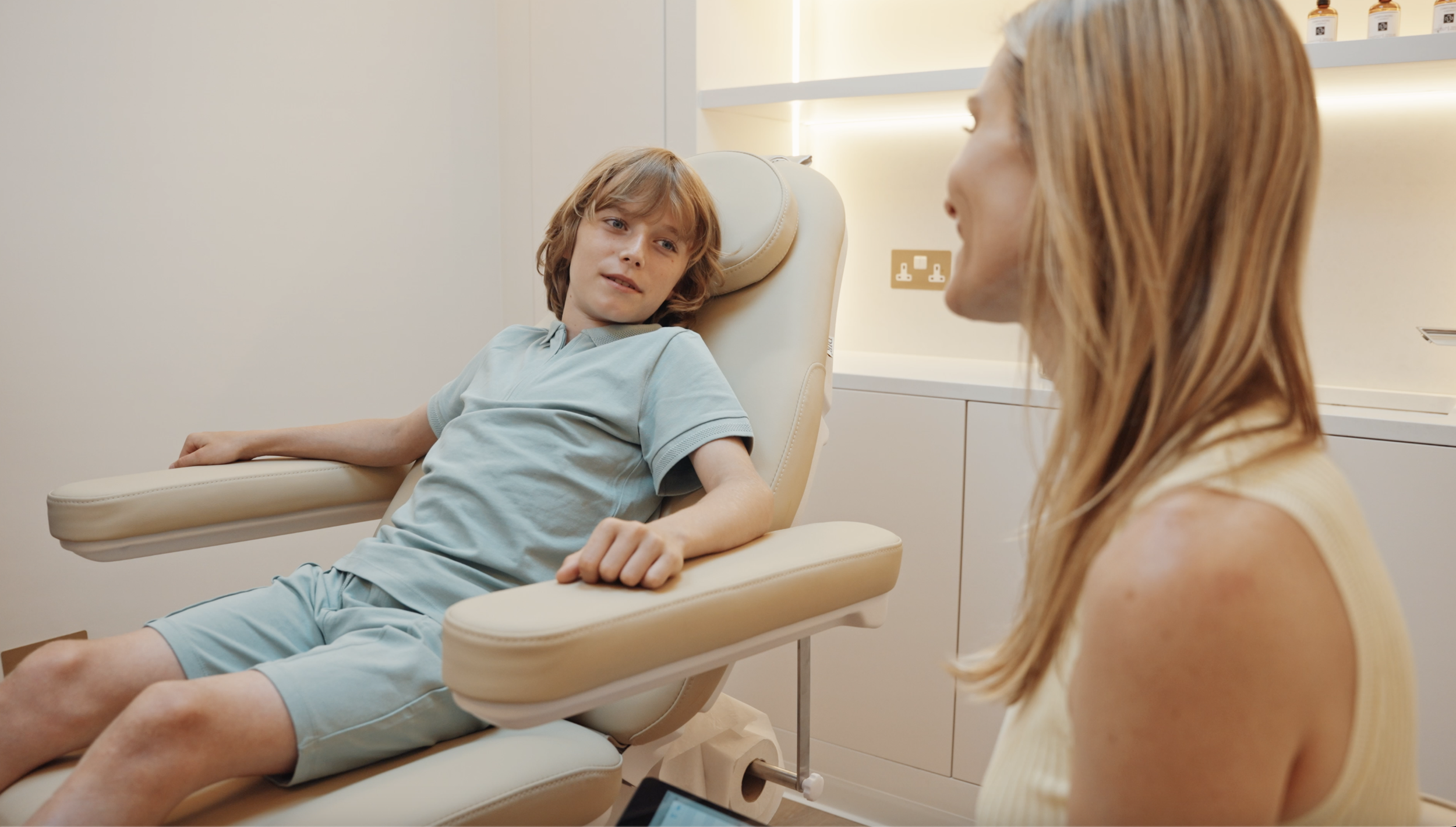 A young boy sitting on a medical examination chair, talking to a woman, in a clinical setting.