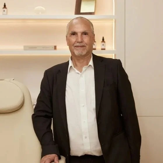 Older man with a beard and bald head wearing a black blazer and white shirt, standing indoors in front of white shelves with decorative items.