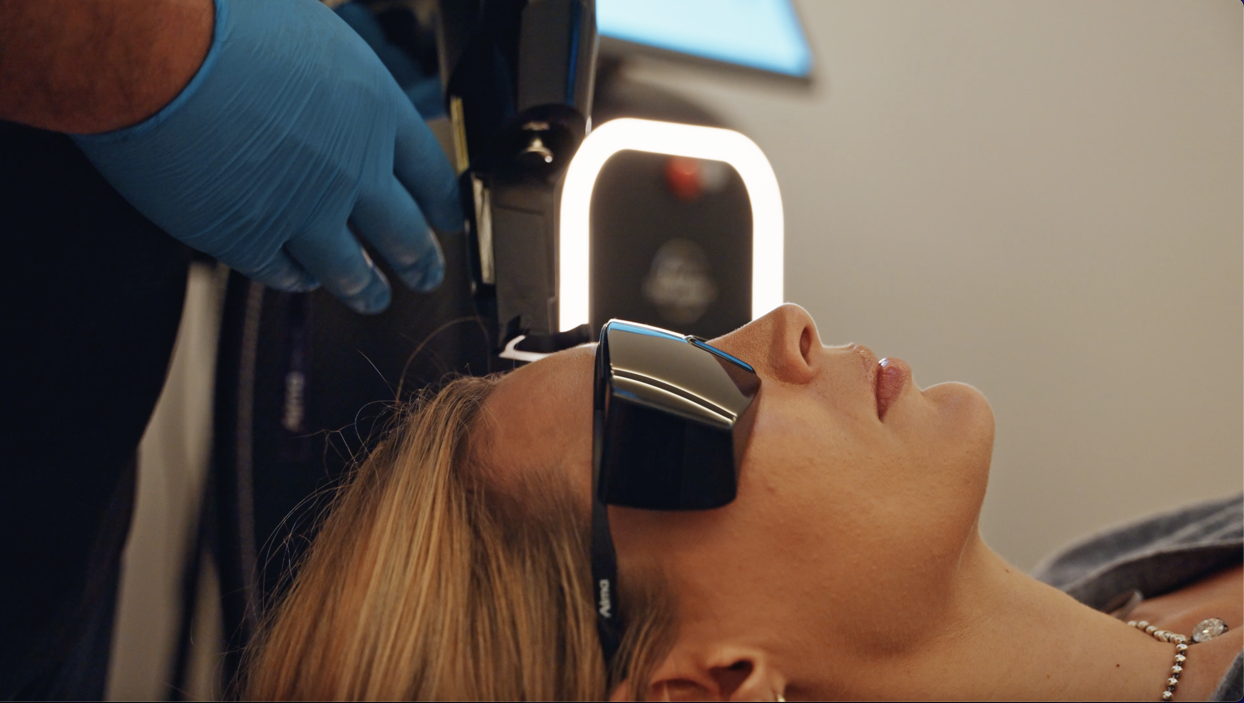 A woman with blonde hair lying back with closed eyes, wearing protective sunglasses, during a facial or skin treatment, with a gloved technician applying a device to her face in a clinic setting.