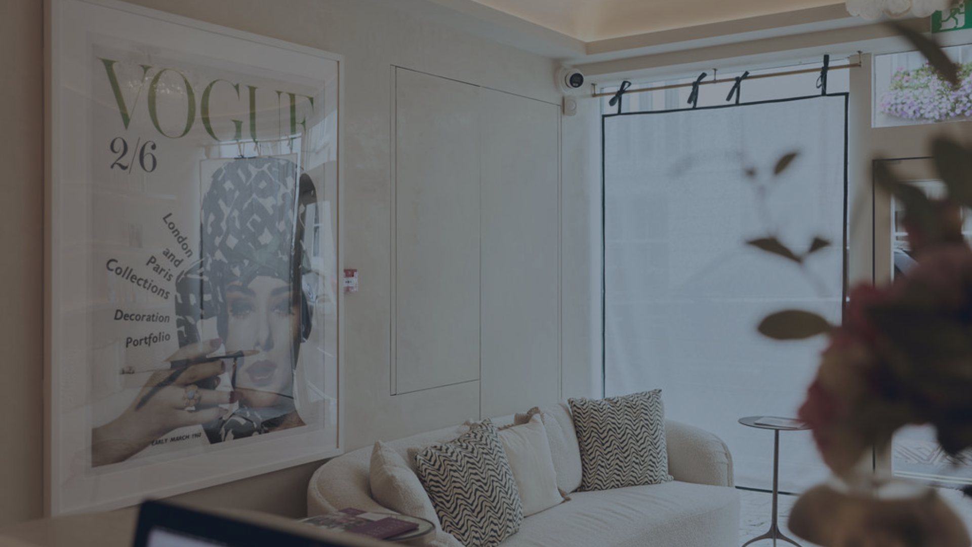 Living room with white brick wall, beige ceiling with exposed beams, black bookshelves, a beige sofa with red and beige pillows, and a black radiator cover with a potted plant on top.