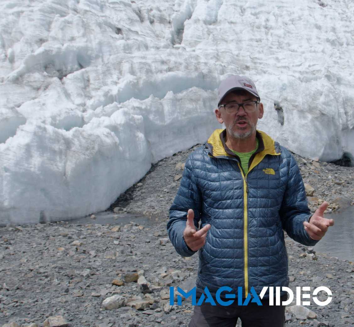 Hombre con chaqueta azul, gorra y gafas, frente a un glaciar, haciendo gestos con las manos.