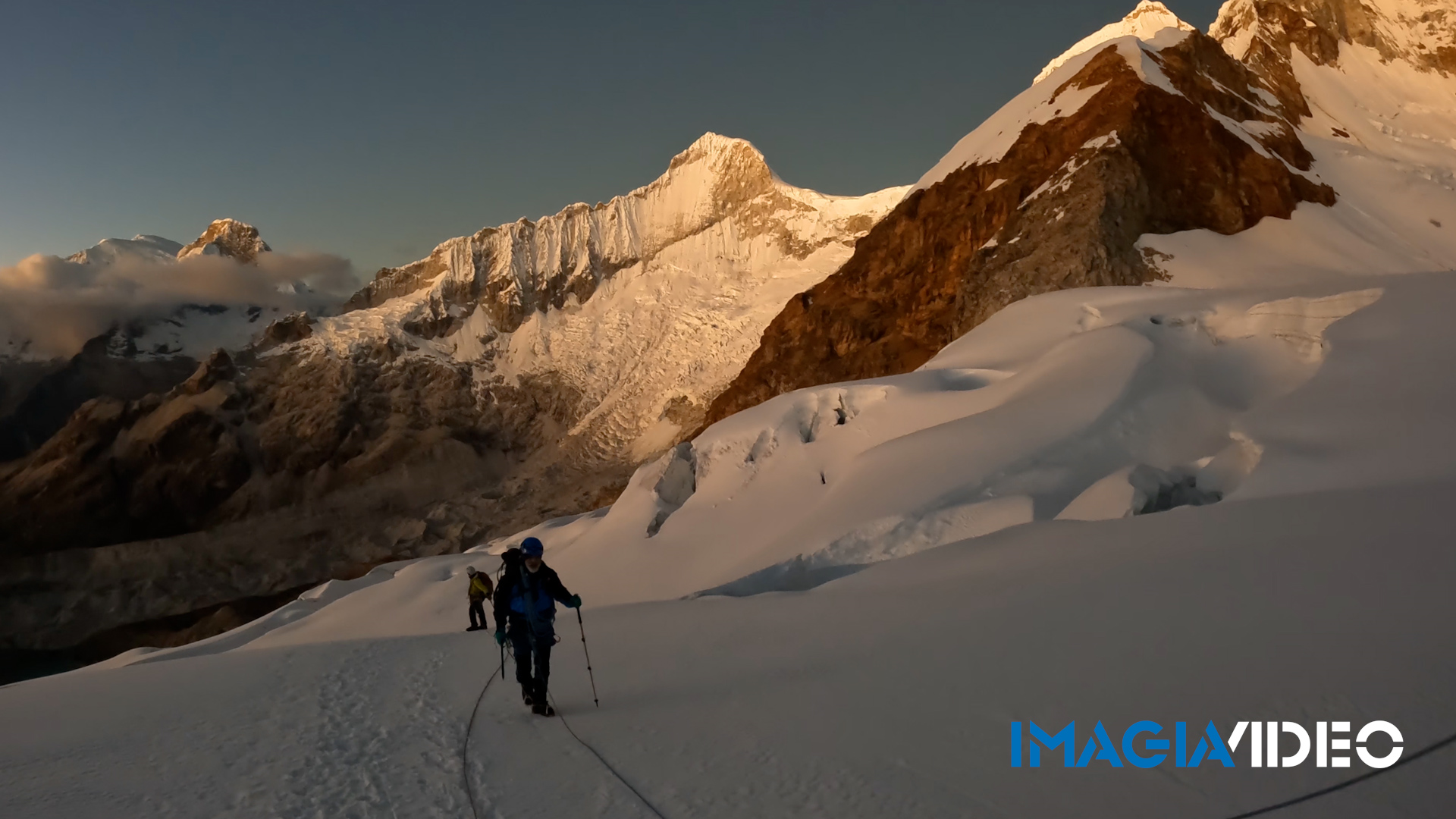 Dos montañistas en una caminata en la nieve en un paisaje montañoso con picos cubiertos de nieve y rocas, al amanecer o atardecer.