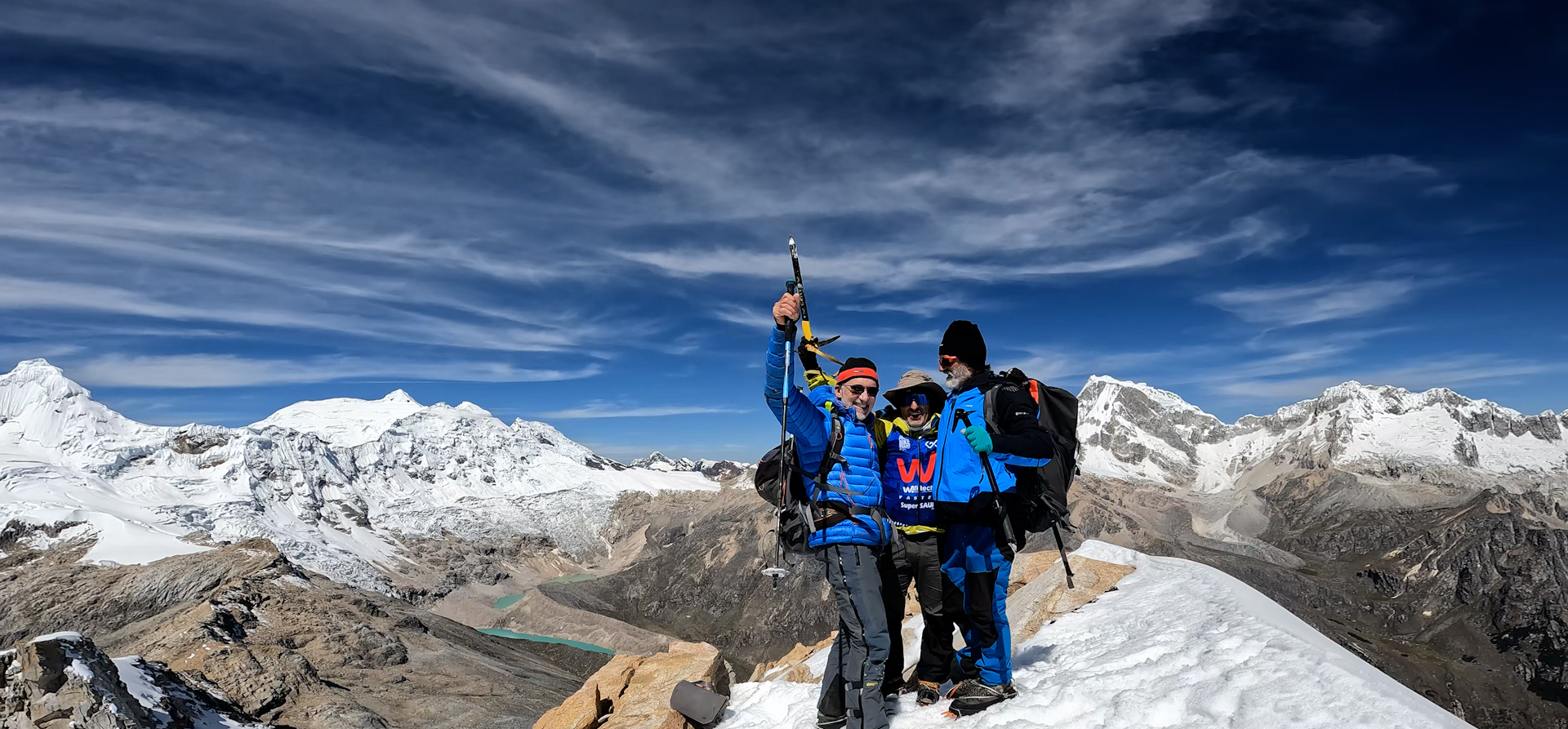 Tres escaladores en la cima de una montaña nevada, celebrando con montañas y un cielo azul con nubes en el fondo.
