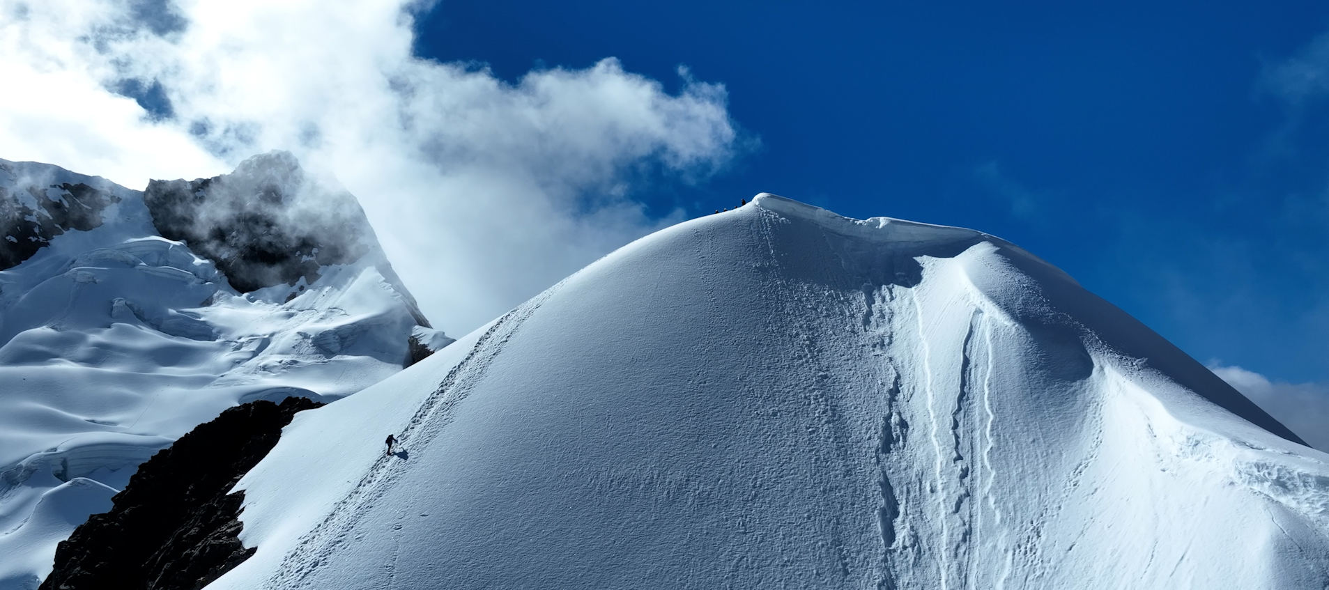 Cima de una montaña nevada con huellas en la nieve y un escalador en la pendiente, cielo azul con nubes