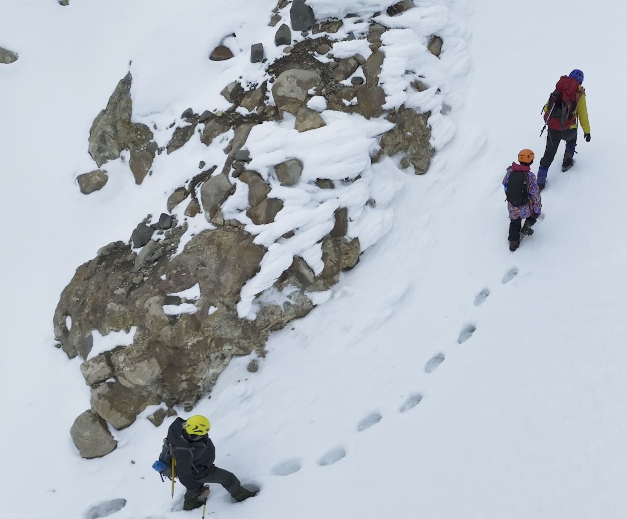 Tres montañistas con equipo de escalada caminando en nieve cerca de formaciones rocosas en una montaña cubierta de nieve.