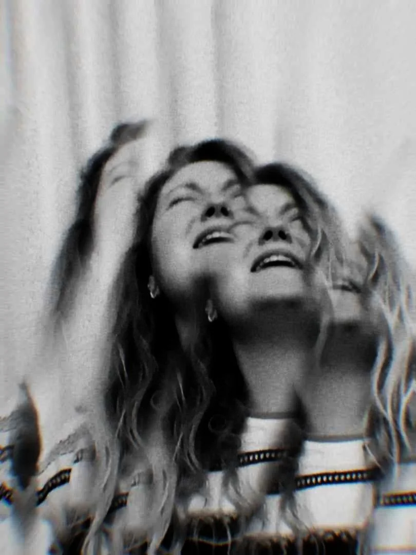 A black and white photo of a white woman has been layered to create a reflective effect. Her head is thrown back and she is singing.