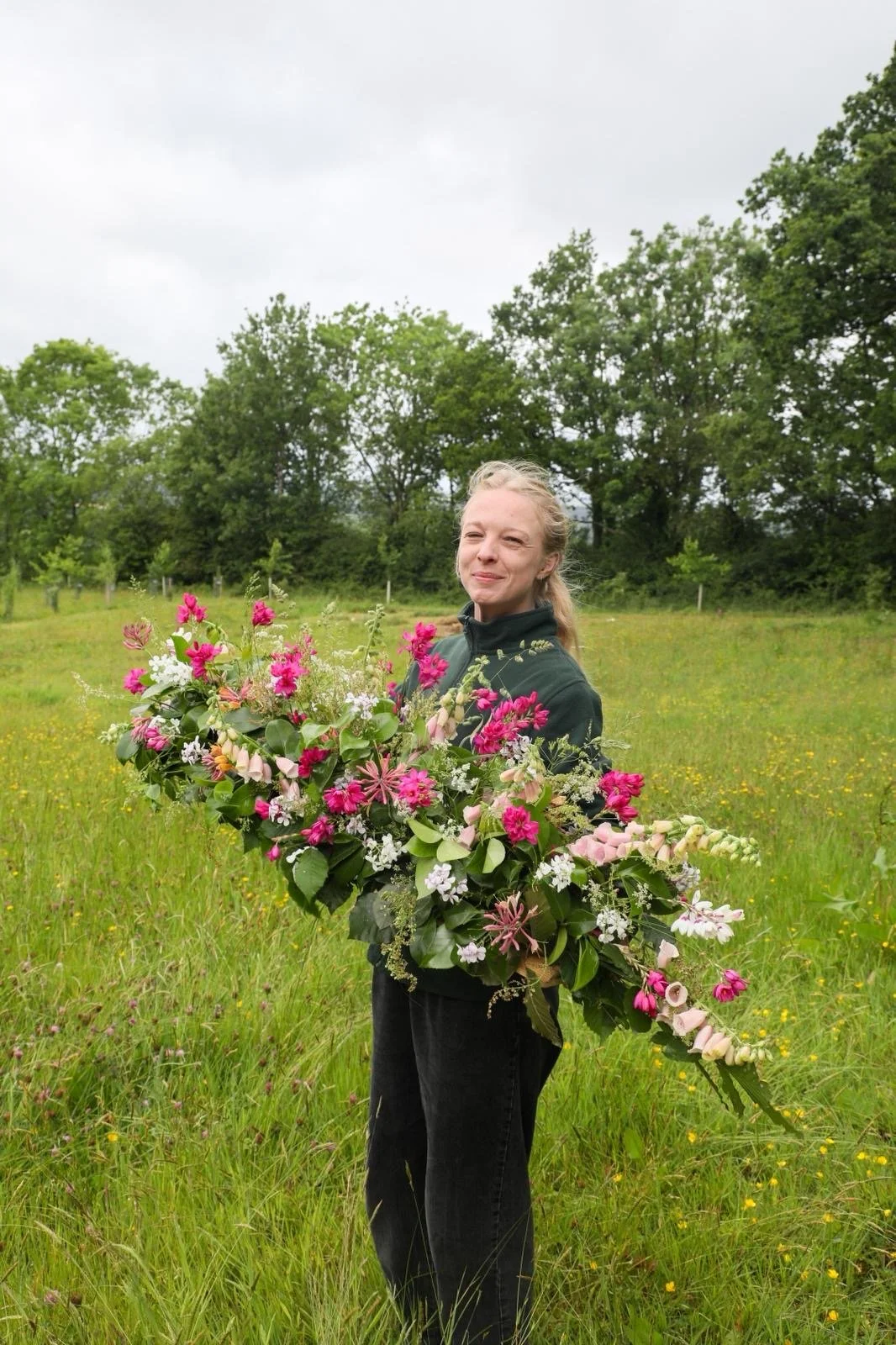 A woman holding a large bouquet of pink, white, and purple flowers in a grassy field with green trees in the background under an overcast sky.