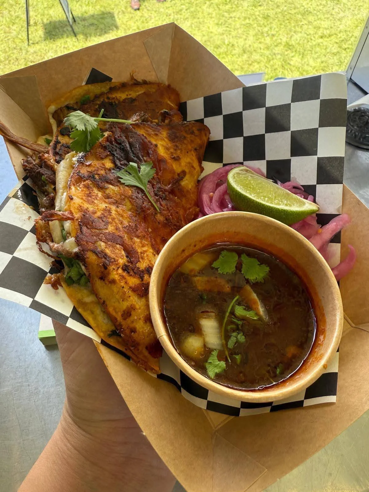 A plate of food containing a grilled meat dish, sliced red onion, a lime wedge, and a bowl of dark broth with herbs.