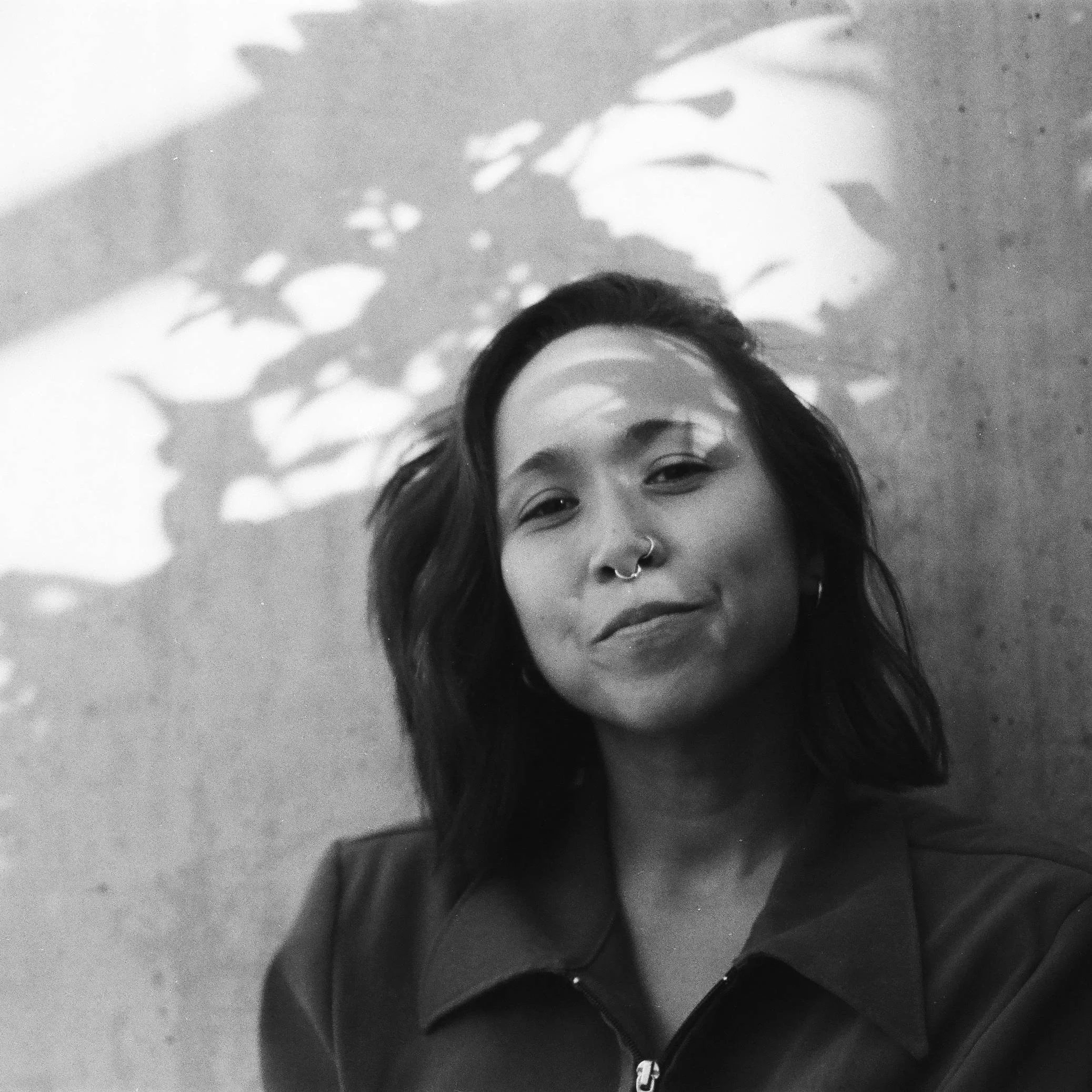 A woman who describes herself as mized east asian heritage wears a nose ring and smiles at the camera. Black and white photo and shadows of plants are on her face.