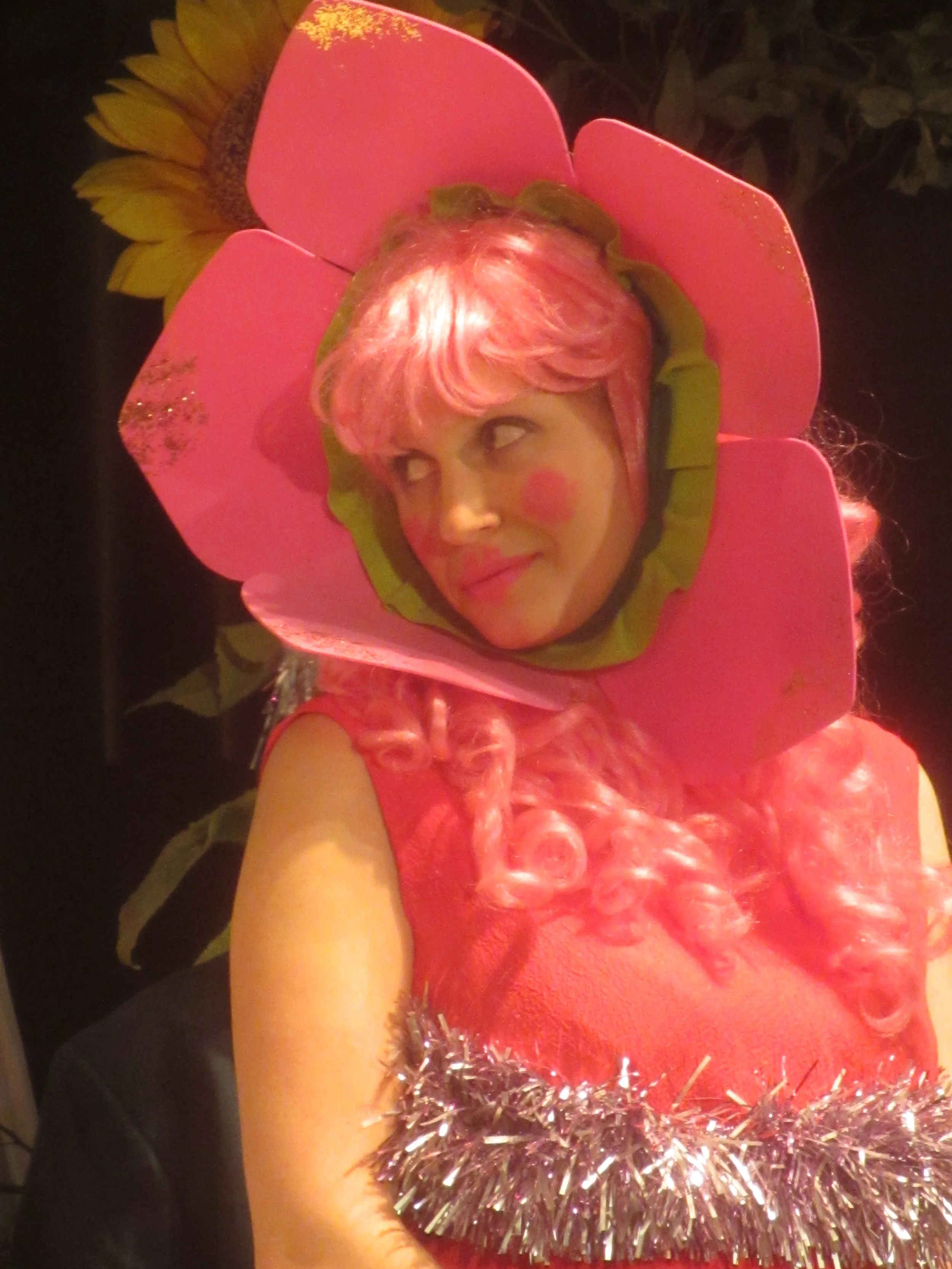 A white woman dressed as a pink flower looks back over her shoulder and smiles softly.