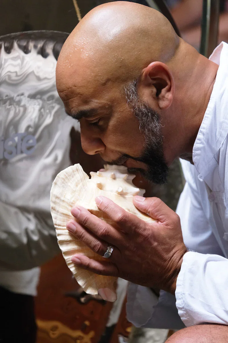 A man with a shaved head and beard is holding a large seashell close to his mouth, appearing to kiss or lick the shell.