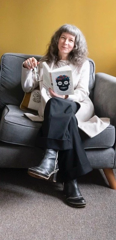 Woman with gray hair sitting on a gray couch, reading a book with a colorful skull cover, against a yellow wall.