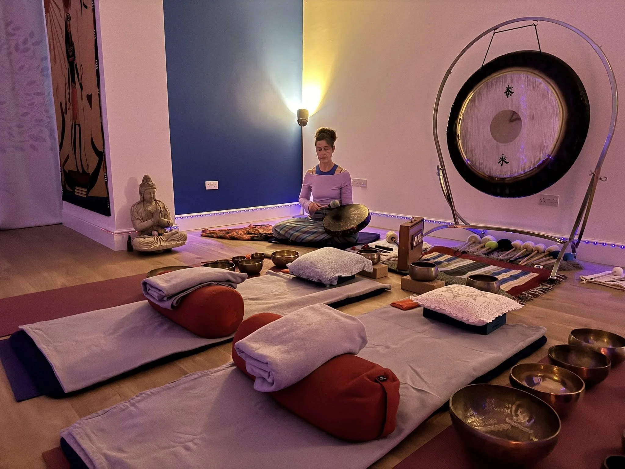 A white woman is seated at the back of a room near a gong, in front are yoga mats. Her eyes are closed and she looks peaceful.