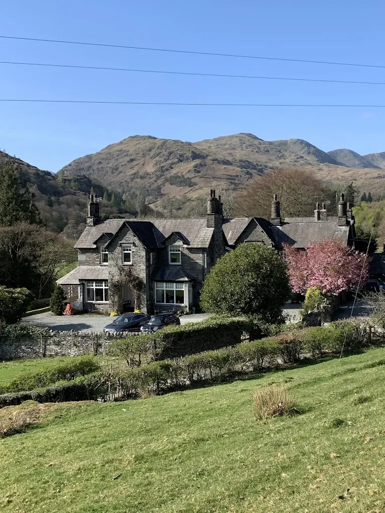 A large stone house with multiple chimneys, surrounded by green trees and shrubs, with mountains in the background under a clear blue sky.
