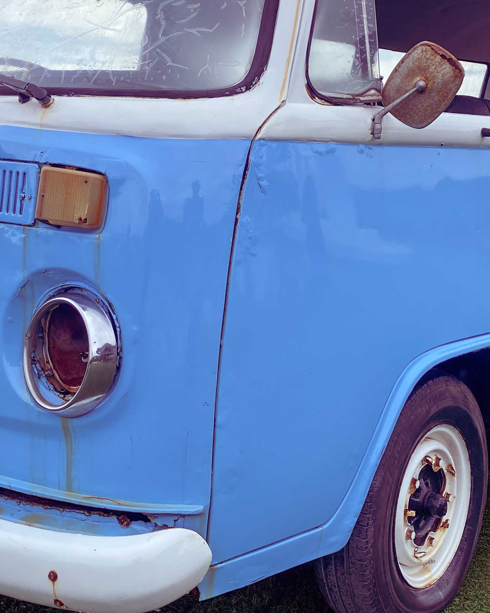 Close-up of vintage blue Volkswagen van with rust spots, cracked windshield, rusty side mirror, and a missing headlight cover.