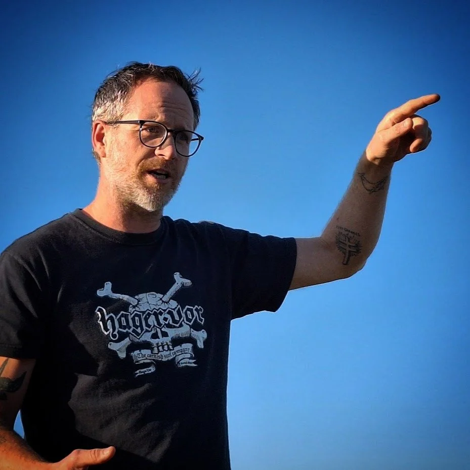 A white man wears a black t shirt in front of a blue sky. He is talking and gesticulating with his left arm.