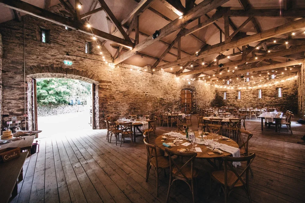 Interior of a large rustic barn with wooden beams and stone walls, string lights hanging from the ceiling, and sunlight coming through an open door to an outdoor area.