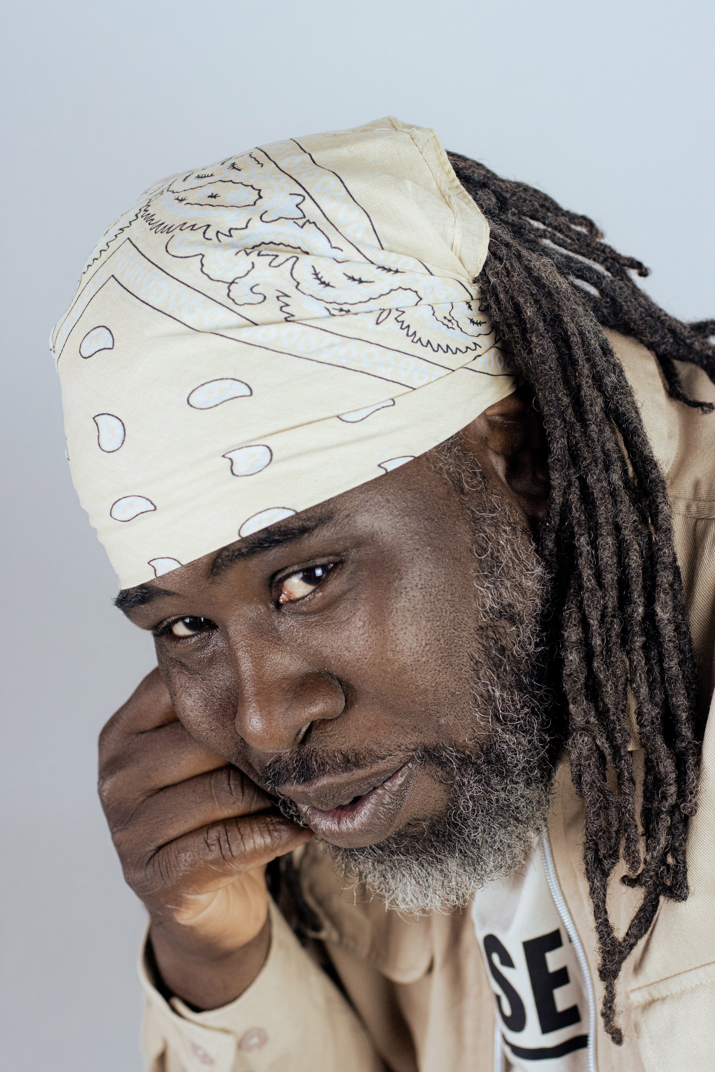 Solo Banton smiles softly at the camera. He is Black and wears a cream bandana over mid length dredlocks, his head is leaning on his hand.