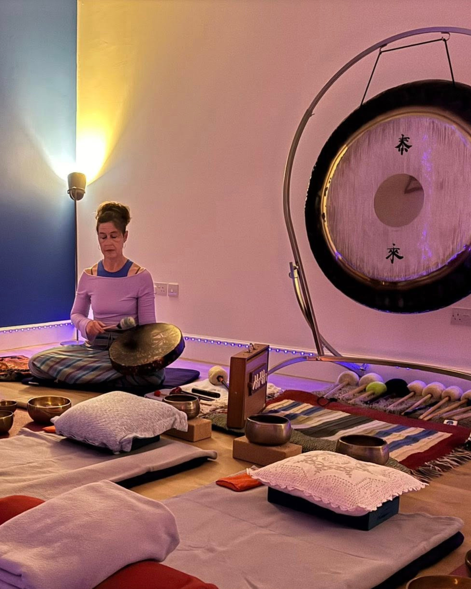 A woman sitting on a cushion with singing bowls and a gong around her, preparing for a sound therapy session in a room with soft pink and blue lighting.