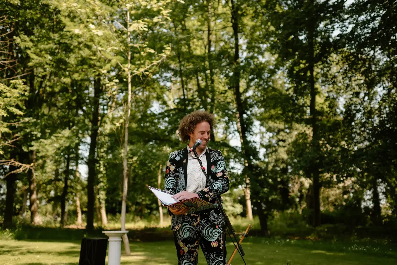 A white man wearing a highly patterned suit is smiling behind a microphone.