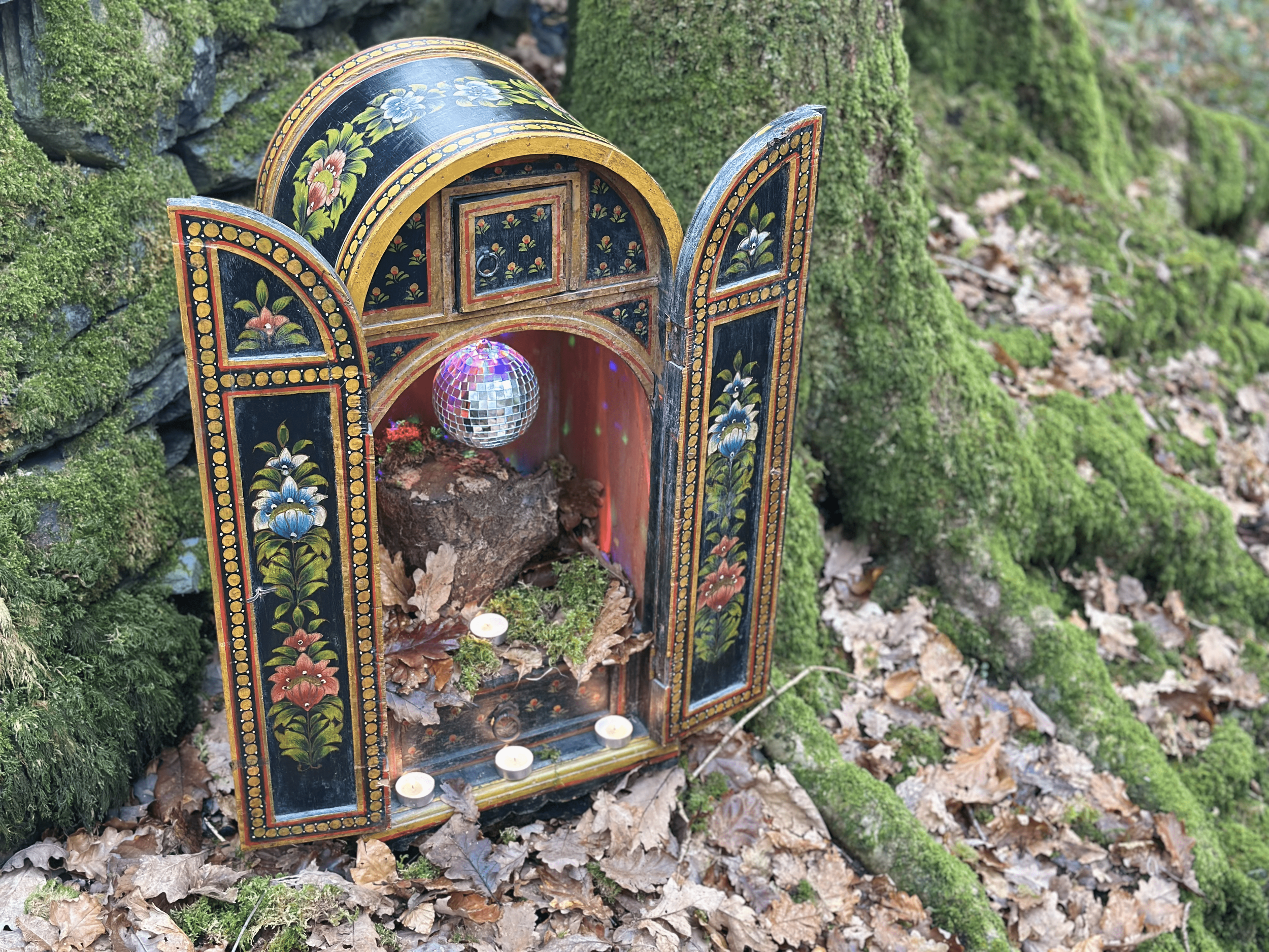 Decorative shrine with floral patterns, a small disco ball, and tealight candles, placed on the ground amidst fallen leaves and moss-covered roots.