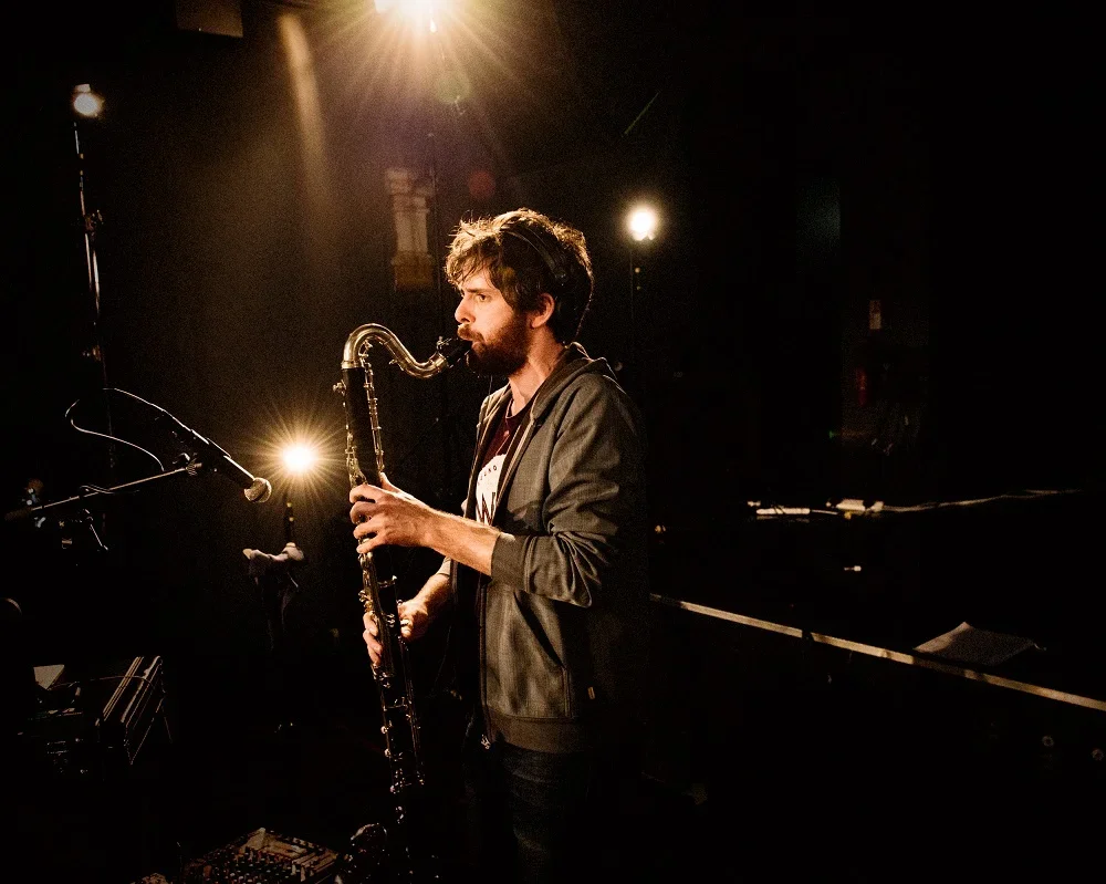 A white man with dark hair and a beard wearing a green jacket playing the clarinet.