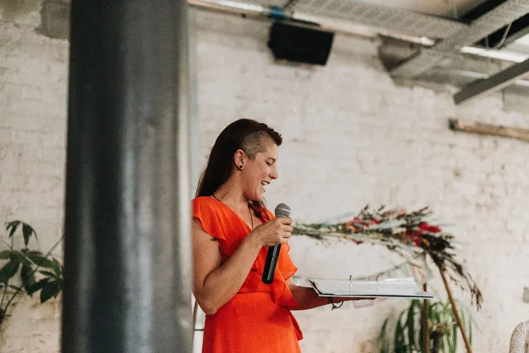 A white woman wears a bright orange dress and is holding a microphone.