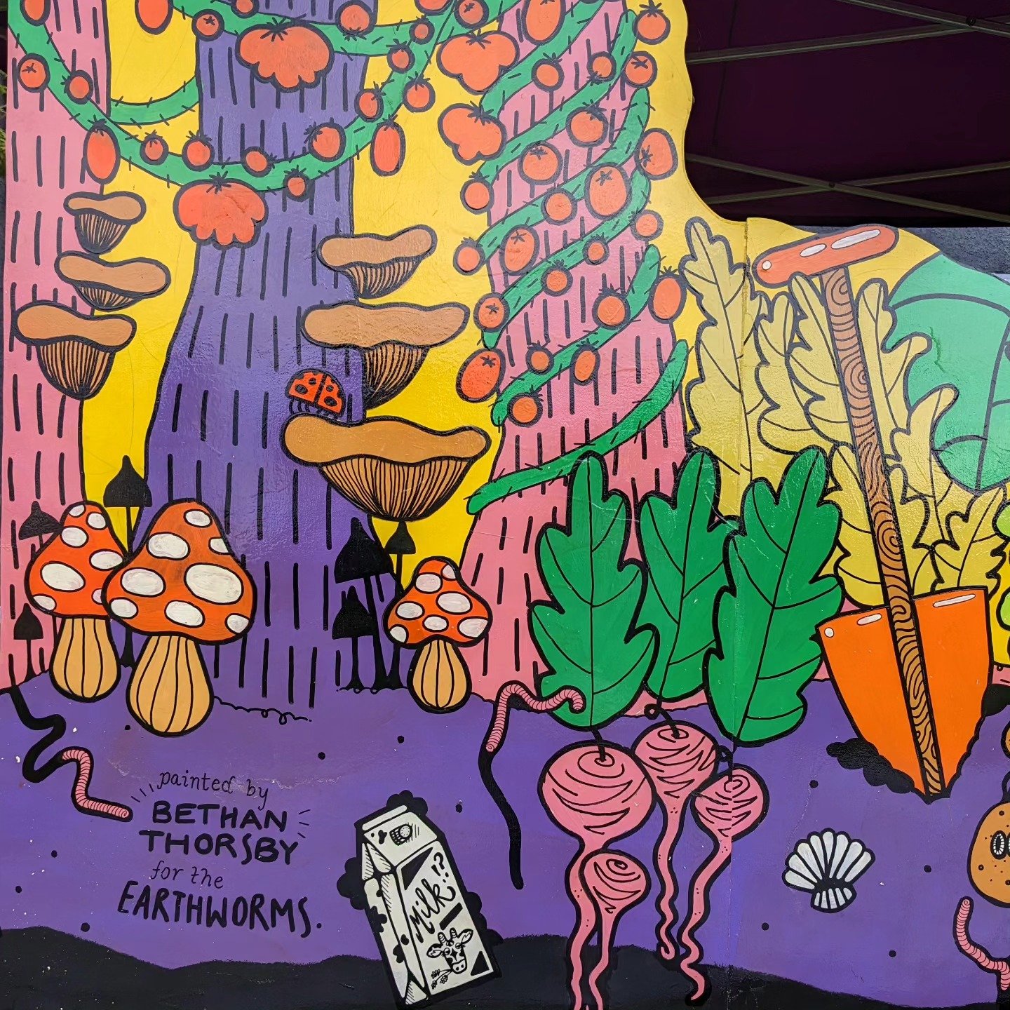 Colorful mural depicting oversized mushrooms, green radishes with pink roots, large green leaves, a packet of milk, a ladybug on a mushroom, a worm, a shell, and a stylized tree trunk with mushrooms and berries, all in a vibrant cartoon style. The mural is signed by Bethan Thorsby for the Earthworms.
