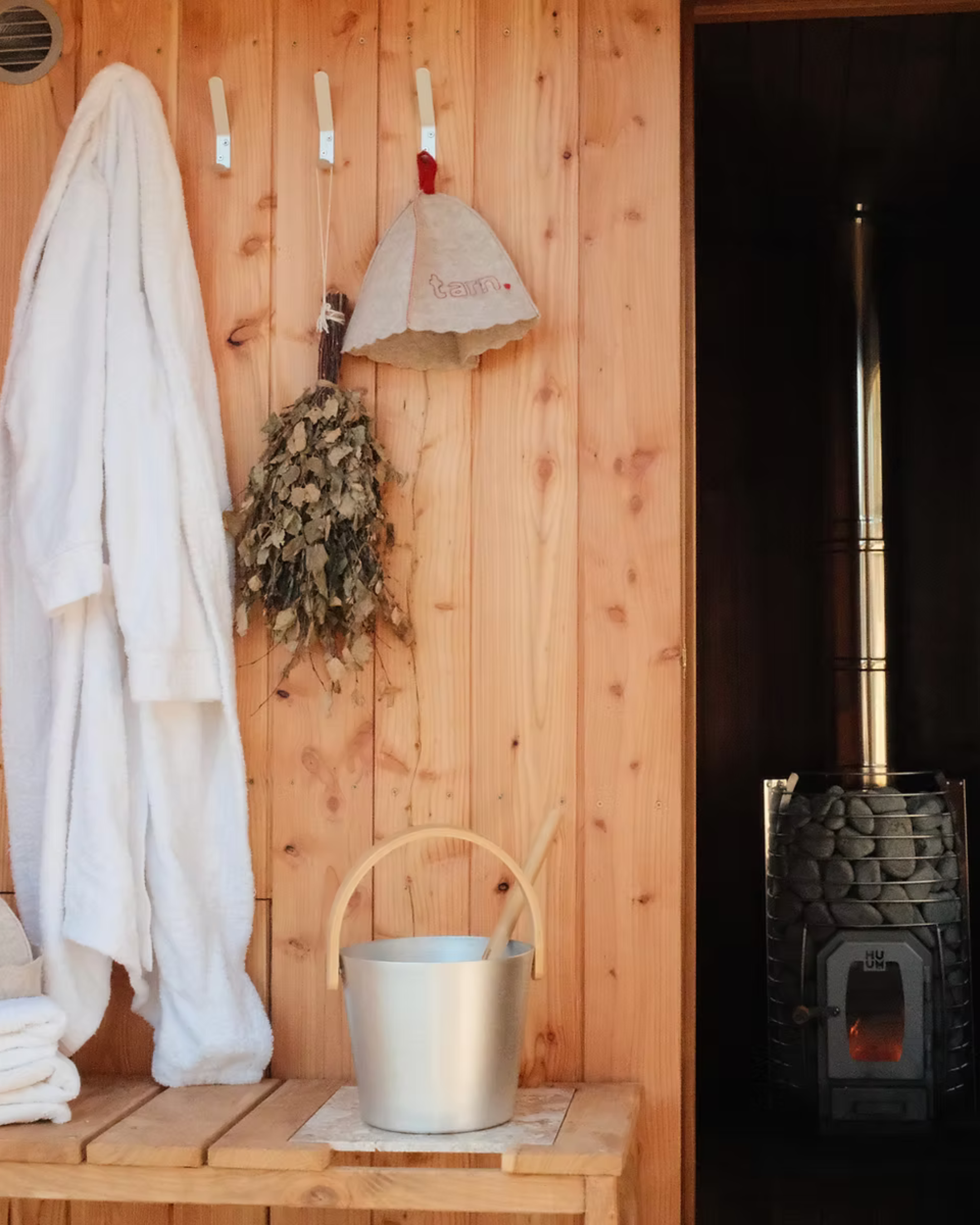A wooden wall with hooks holding a white bathrobe, a bundle of dried herbs, a beige cloth bag, and a paper bag with a red drawstring. A wooden table below holds a metal bucket with a wooden handle and a ladle. To the right, a black stove with rocks and a pipe for heating the room.