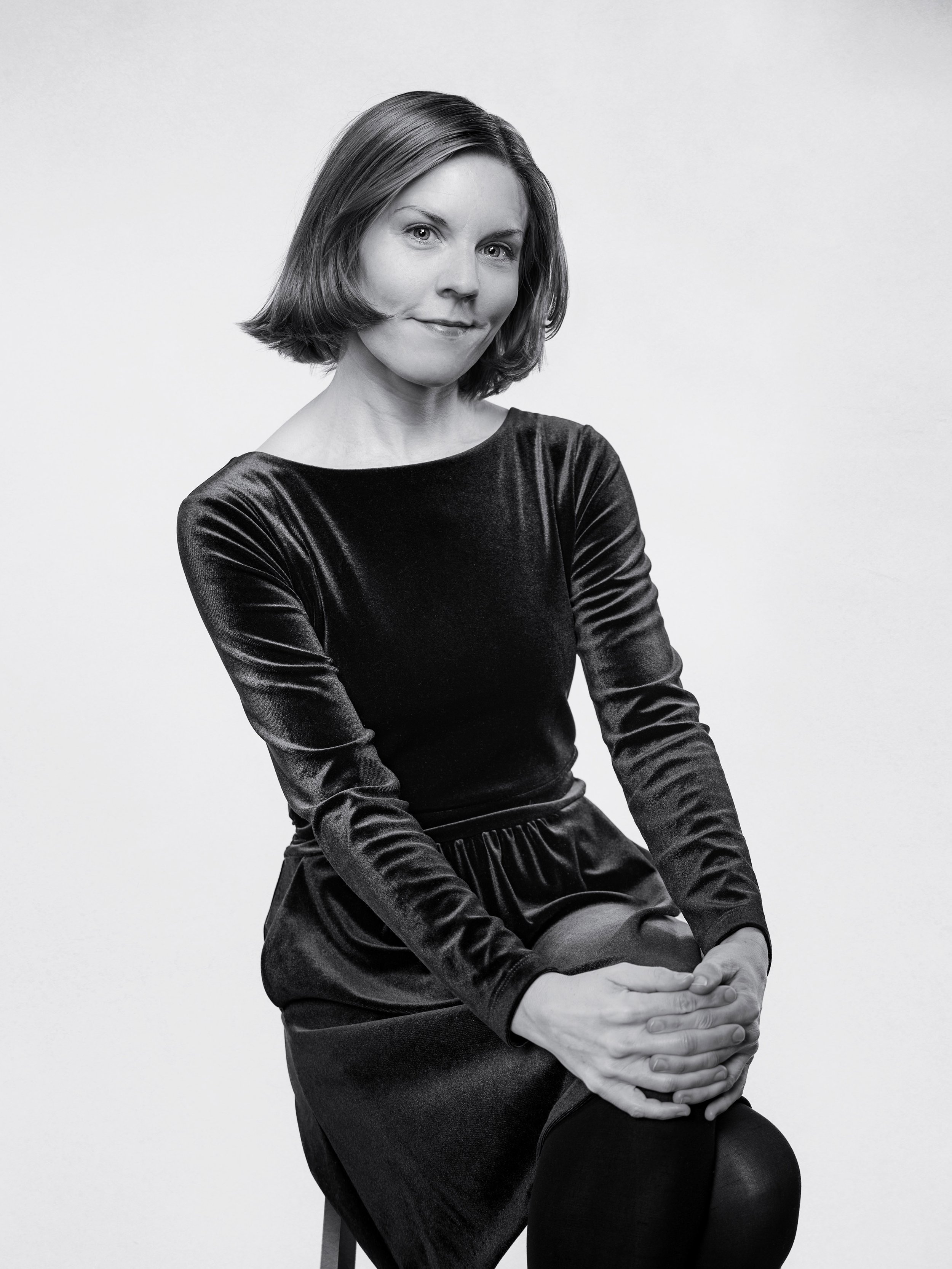 a white woman with a bob, sits on a stool and smiles at the camera. She is wearing a black velvet dress.
