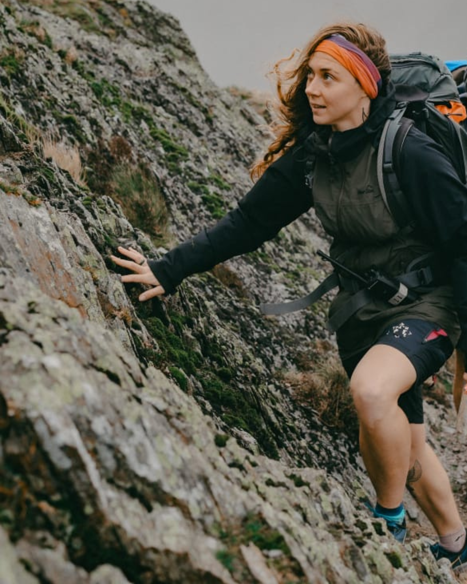 A woman climbing a rugged, moss-covered rock face outdoors, wearing outdoor gear and carrying a backpack.