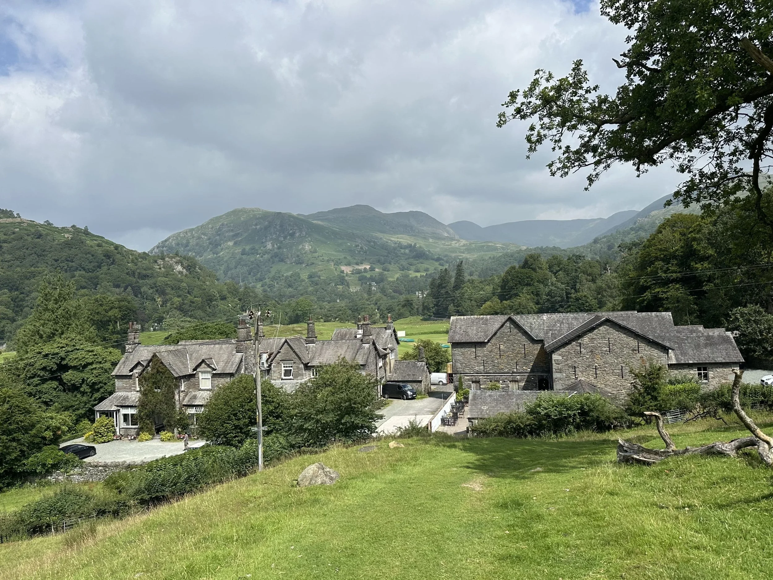 Scenic view of a farm with stone houses, surrounded by green hills and mountains under cloudy sky in Rydal, Lake District.