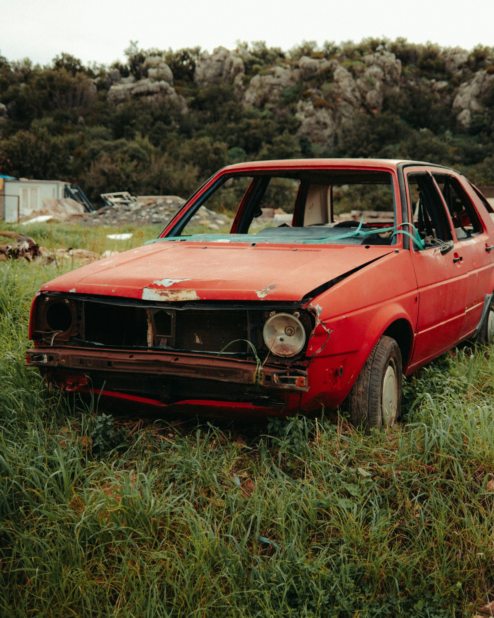An abandoned red car with missing front panel, no headlights, and overgrown grass surrounding it, set outdoors with rocky hills in the background.