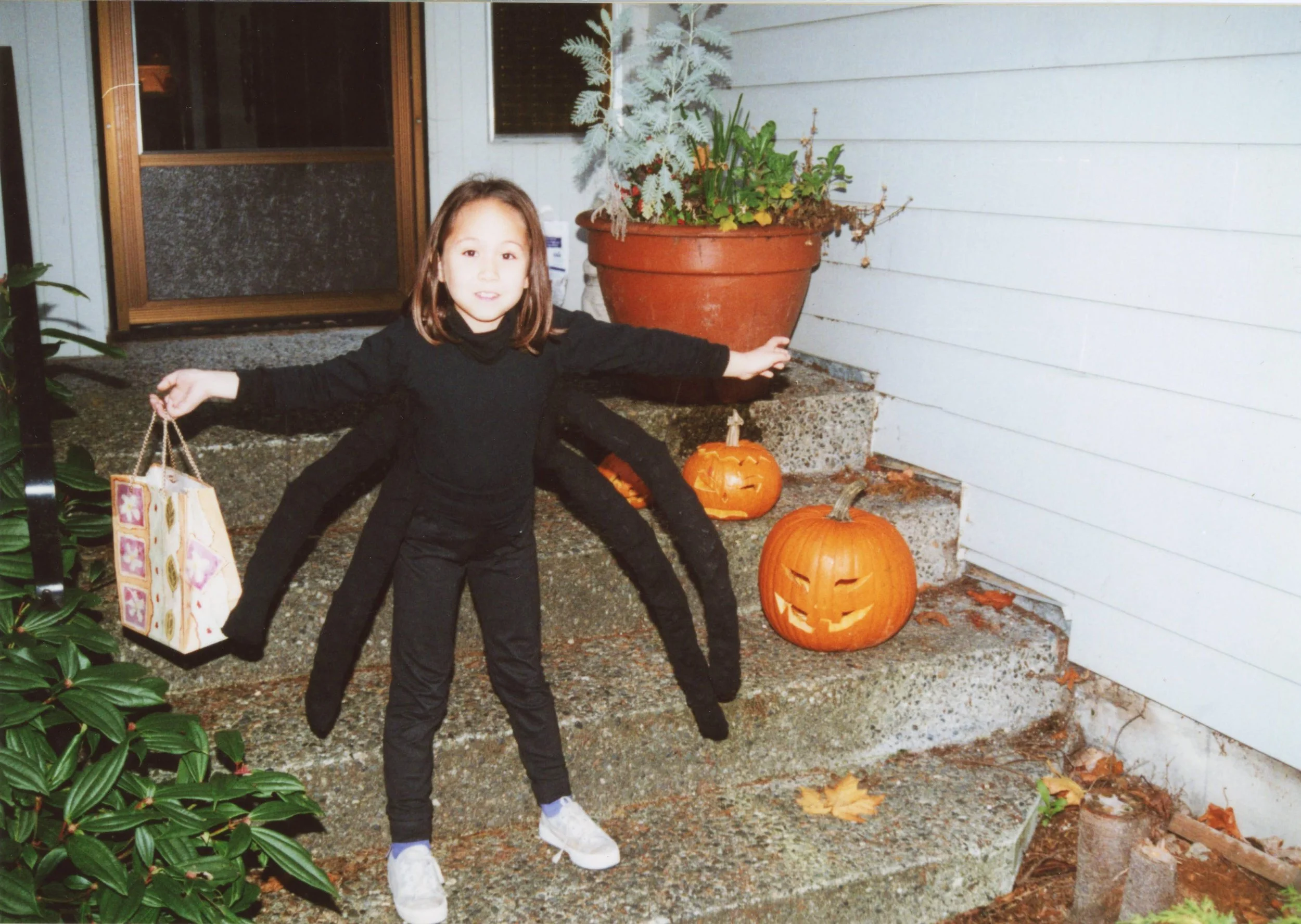 A young girl with mixed heritage (Chinese American) wears a homemade halloween spider costume. She stands on steps holding a colourful paperbag, two Jack O Lanterns can be seen on the steps.
