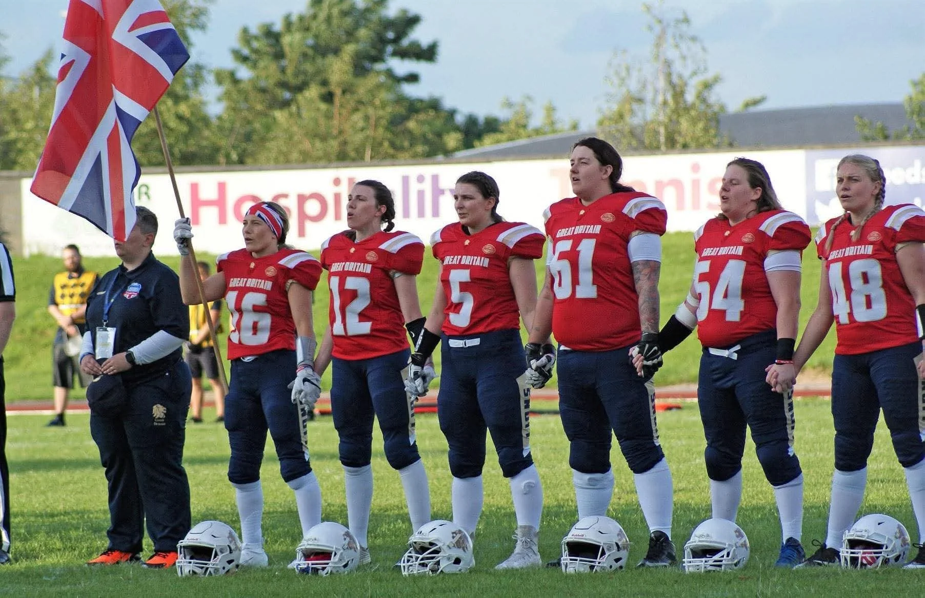 women's American football team standing in a line holding hands before a game, with a person in uniform holding a British flag. he players are wearing red jerseys with white numbers, blue pants, and white knee-high socks, with helmets placed on the ground in front of them.