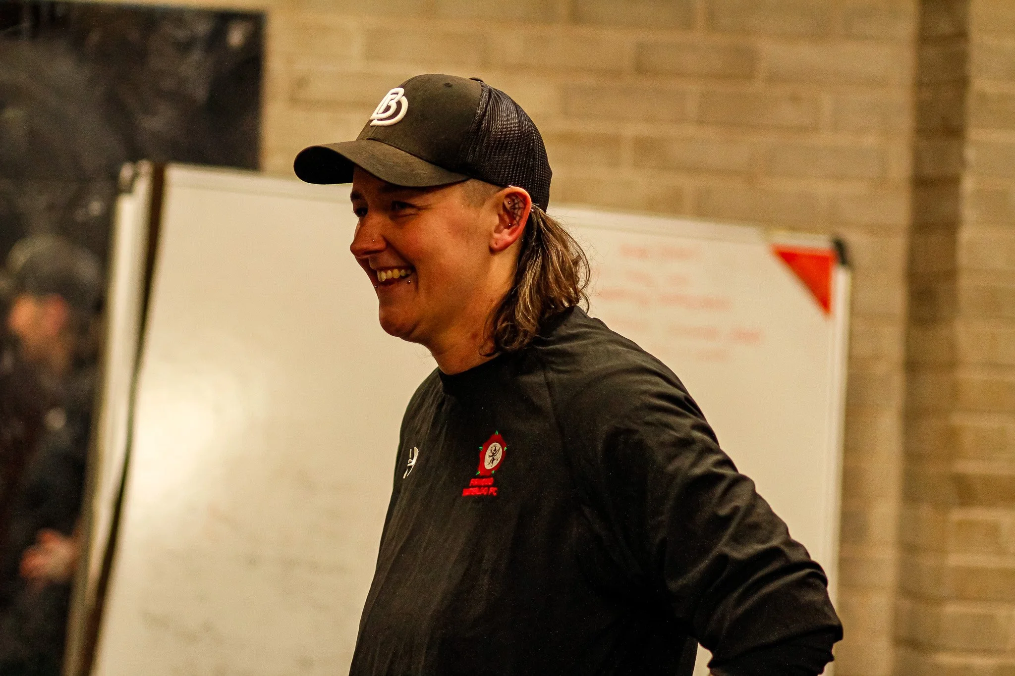 smiling female coach with long hair wearing a baseball cap and black jacket, in an indoor setting.