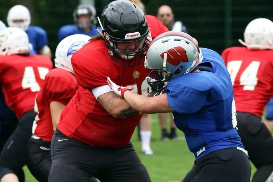 two American football players, one in a red jersey and the other in a blue jersey, engage in a blocking drill during practice on a football field.