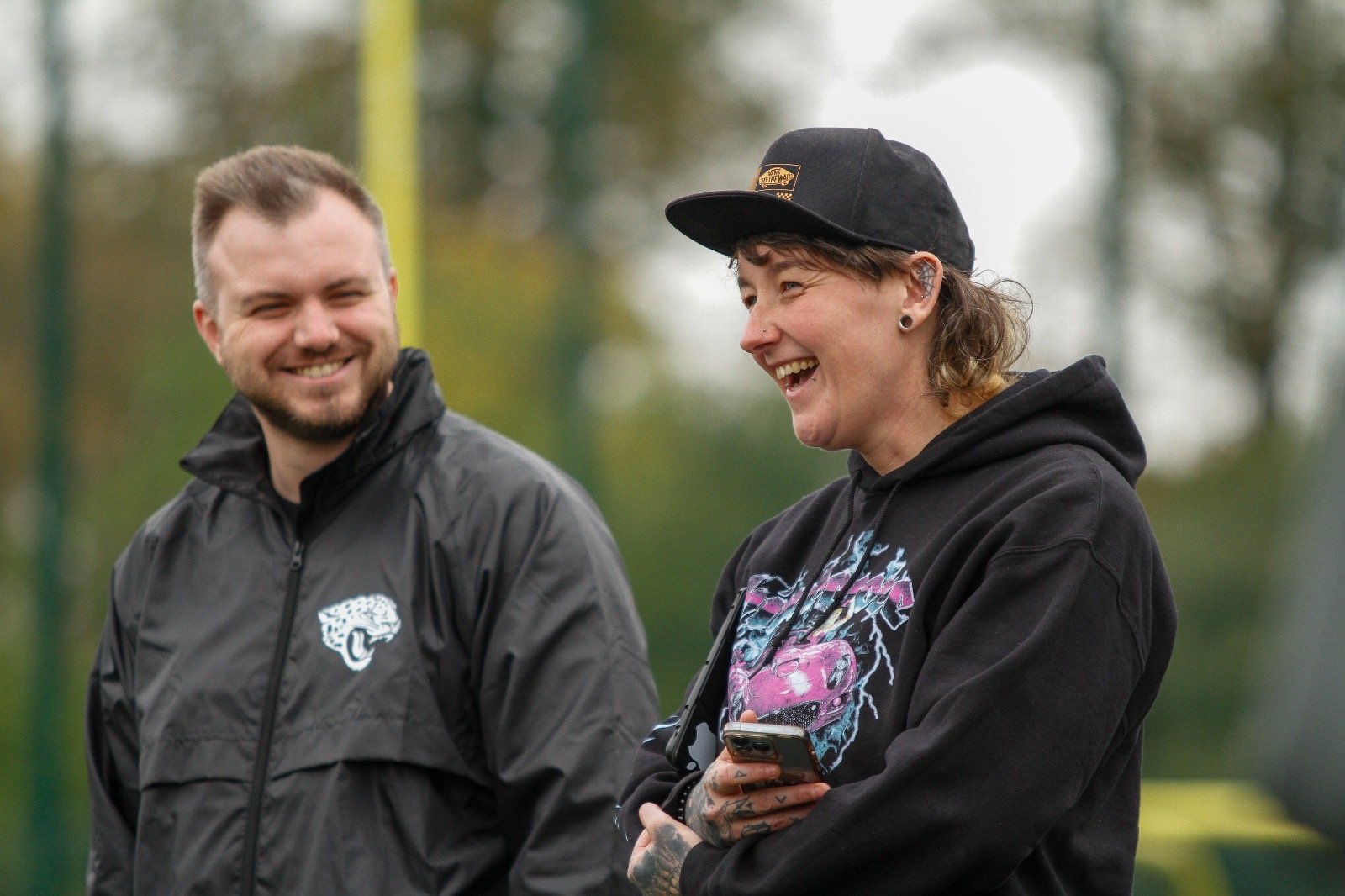 two people smiling and talking outdoors, one wearing a black jacket with a jaguar logo, the other in a black hoodie with a colourful graphic, holding a smartphone.