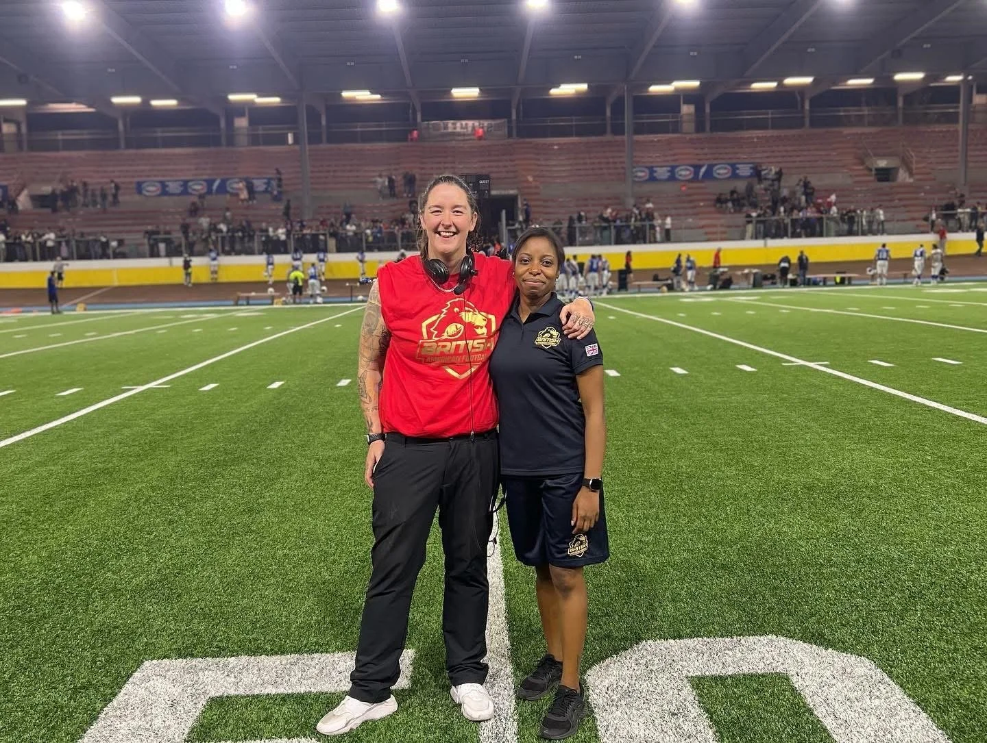 two females standing on a football field inside a stadium, smiling and posing for a photo. one woman is wearing a red sports jersey and the other is wearing a navy blue sports shirt.